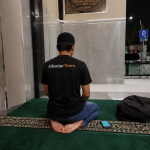 Ilustrasi musafir modern sedang salat qashar di musala rest area malam hari sambil membawa tas perjalanan dan ponsel.