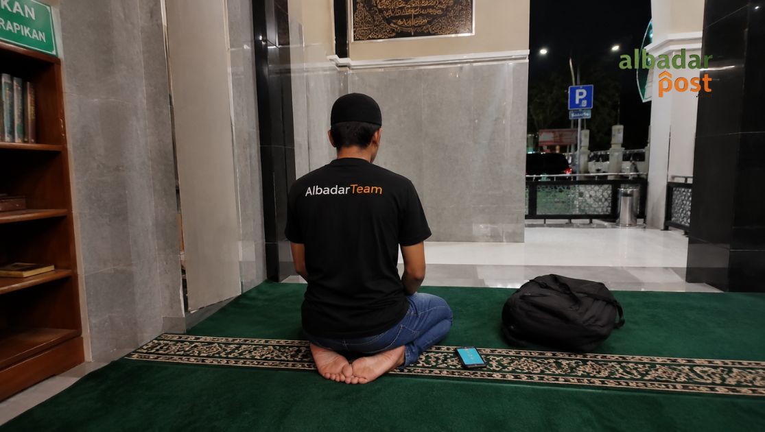 Ilustrasi musafir modern sedang salat qashar di musala rest area malam hari sambil membawa tas perjalanan dan ponsel.