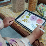 Seorang ilustrator digital menggambar bunga menggunakan tablet dan stylus di meja rumah sederhana dengan suasana hangat.